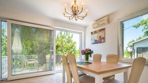 A dining area with a wooden table and six chairs, large windows with blinds, a chandelier, and an air conditioning unit. - Vacation home Vesna by Interhome (Krk/Njivice)