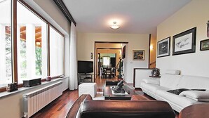 A spacious living room with a white sofa, a black leather chair, and a television on a stand. There is a large window with curtains, a radiator, and a dining area in the background. - Vacation home Vesna by Interhome (Buzet)