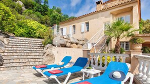 A poolside area with lounge chairs, a stone wall, and a building with a balcony. - Bella Vista by Interhome (Calonge)