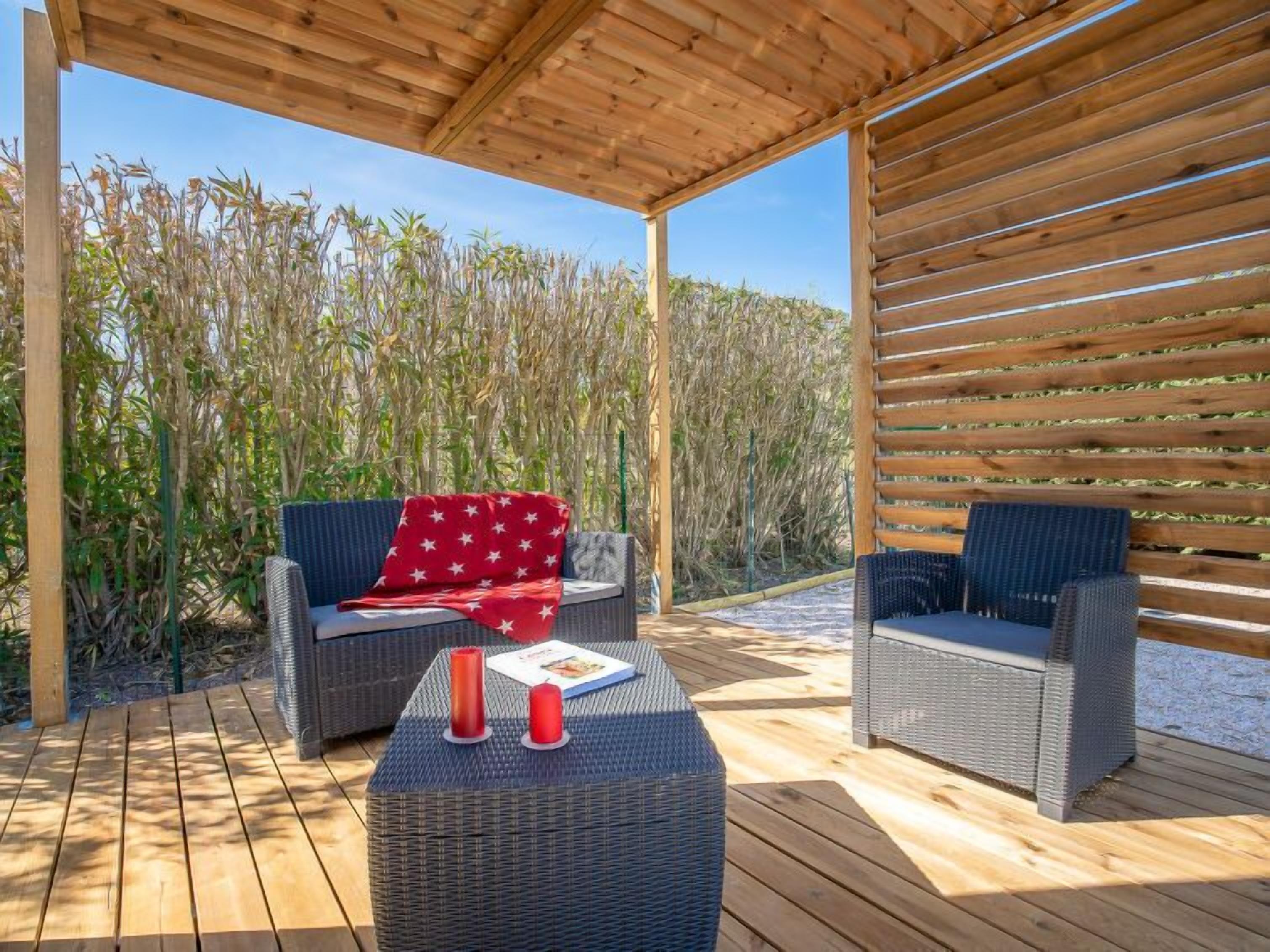 Een terras buiten met rieten meubels, een houten tafel en een rood kussen met sterrenprint.