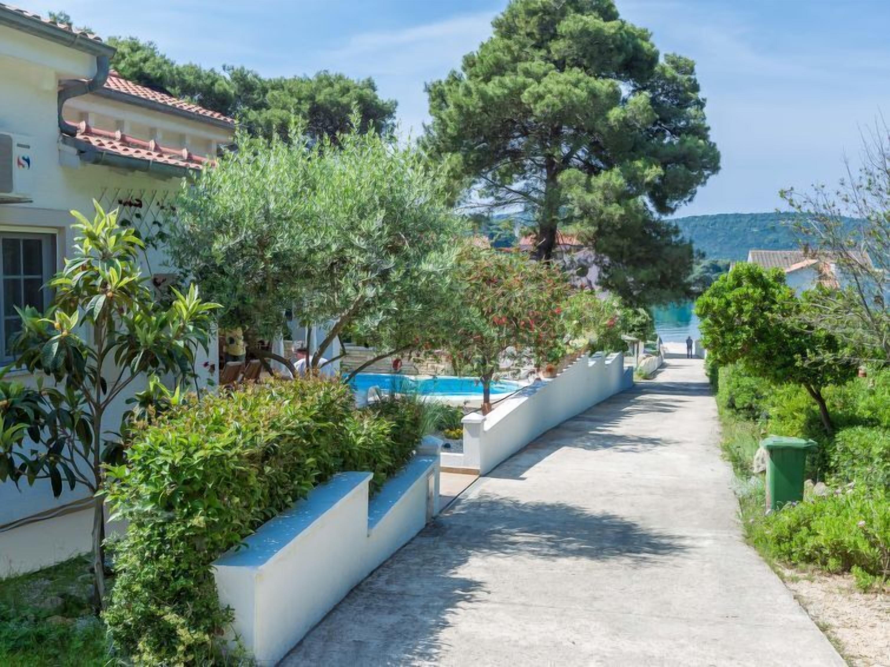 Un chemin mène à un espace piscine entouré de verdure et à un bâtiment doté d’un toit recouvert de tuiles.