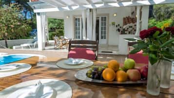 An outdoor dining area with a wooden table, plates, cutlery, and a fruit platter.