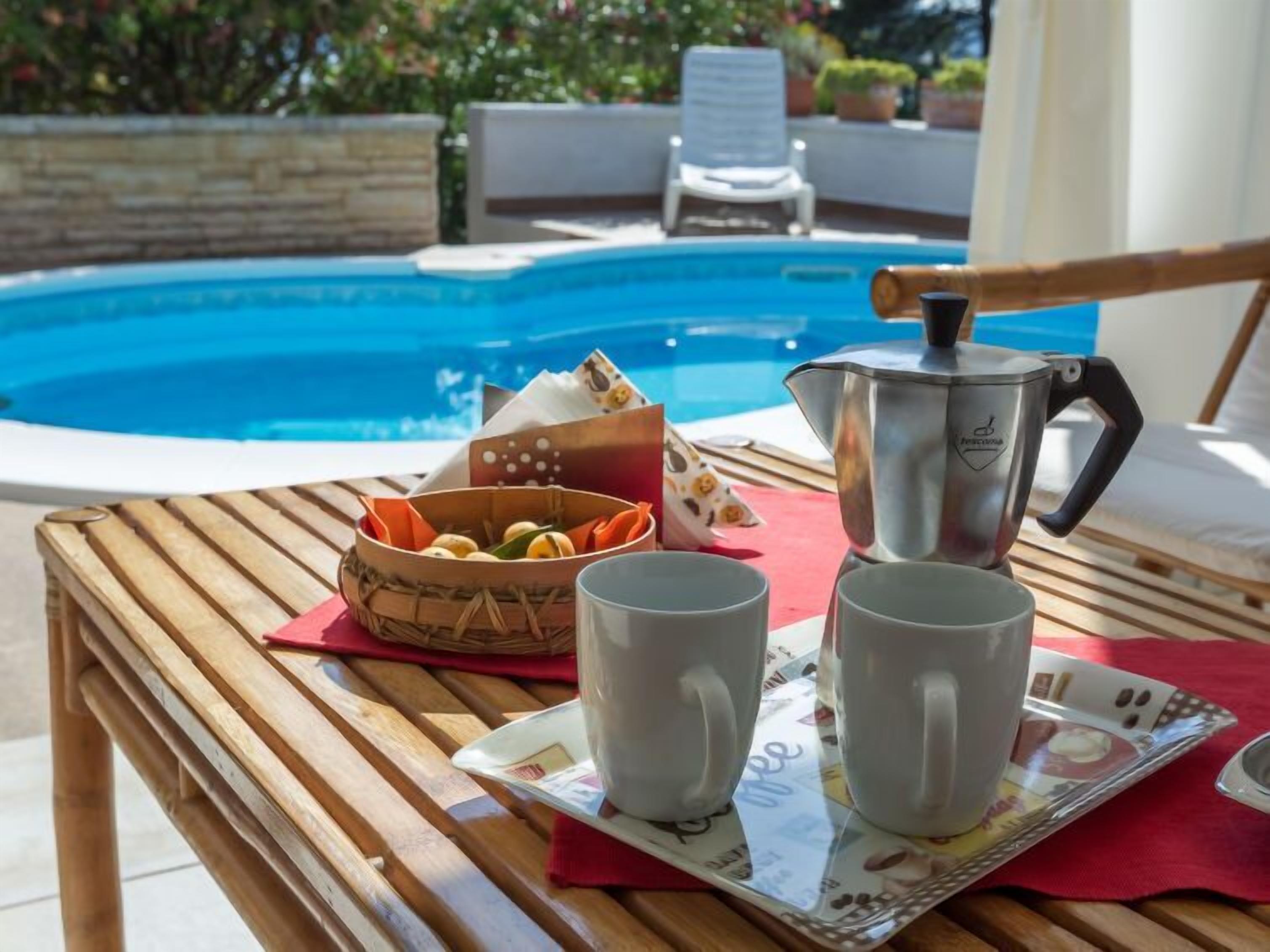 Une table en bois avec un plateau, deux tasses, un panier de fruits et une cafetière, le tout placé près d’une piscine.