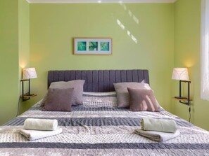 Una cama bien hecha con una colcha gris y blanca estampada, dos almohadas y dos toallas dobladas sobre una pared verde con un cuadro enmarcado arriba.