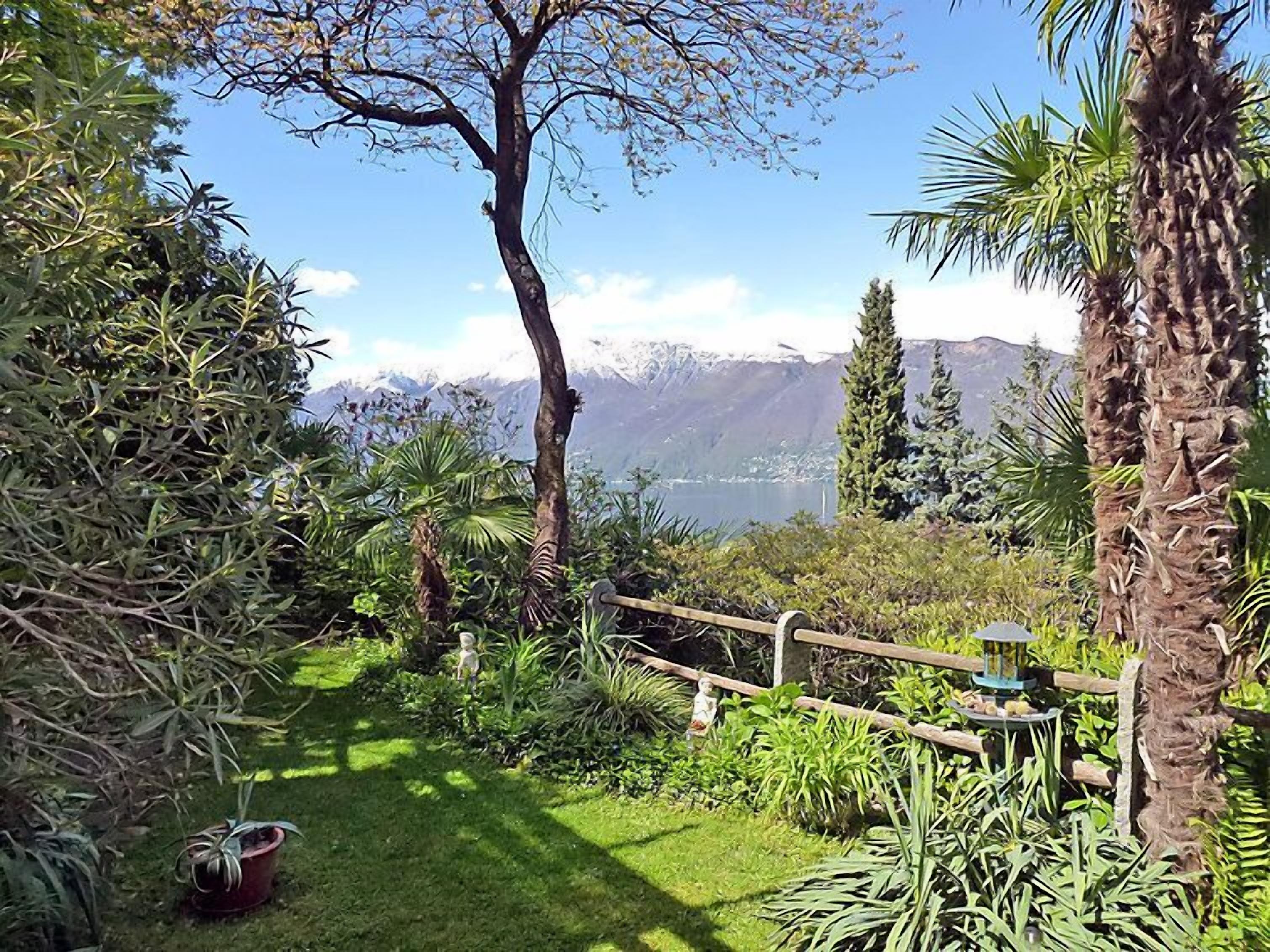 A garden with a wooden fence, various plants, a small statue, and a view of mountains and a lake.