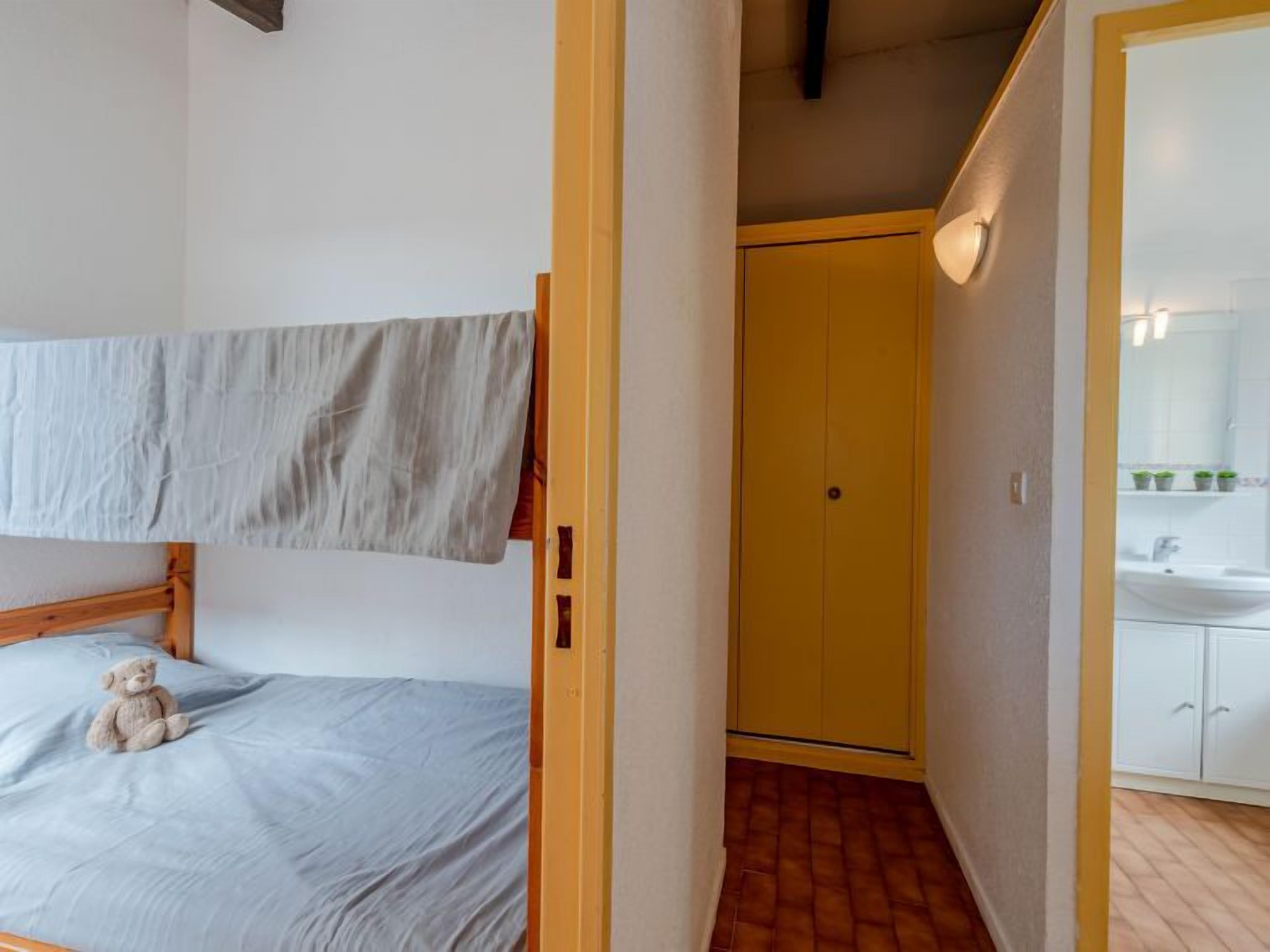 Une chambre avec un lit superposé, un petit ours en peluche sur le lit, une porte jaune, une salle de bain avec un lavabo et un miroir, et un sol carrelé.