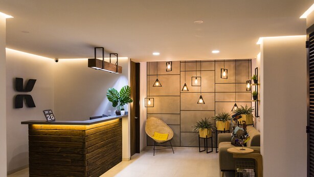 A reception area with a wooden reception desk, a wicker chair, and a wall with geometric patterns and pendant lights. - Kapital Suites (Pereira)