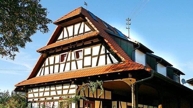Une maison traditionnelle à colombages, dotée d’un toit à forte pente, d’une cheminée bien visible et d’un porche à l’avant, avec des chaises et une table.