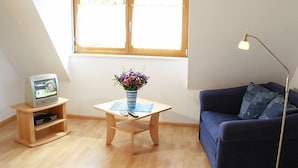 Une pièce avec un plancher en bois, un canapé bleu, une petite table sur laquelle est posé un vase de fleurs, un téléviseur sur un meuble en bois et une lampe sur pied.