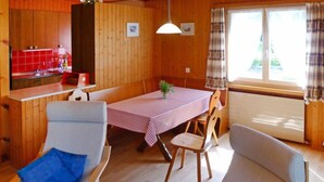 A cozy room with wooden walls, a dining table with a checkered tablecloth, two chairs with blue cushions, a hanging light, and a window with curtains. - Untertal by Interhome (Elm)