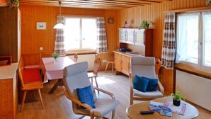 A cozy room with wooden walls and floors, a dining table with a checkered tablecloth, two armchairs with cushions, a small cabinet, and a window with curtains. - Untertal by Interhome (Elm)