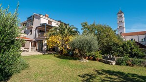 Ein modernes, zweistöckiges Haus mit Balkon, umgeben von einem gepflegten Garten mit verschiedenen Pflanzen und einer Steinmauer.