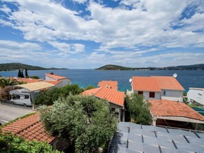 Un quartier résidentiel en bord de mer, avec des toits recouverts de tuiles rouges, un fourgon blanc et une vue dégagée sur la mer et les collines lointaines.