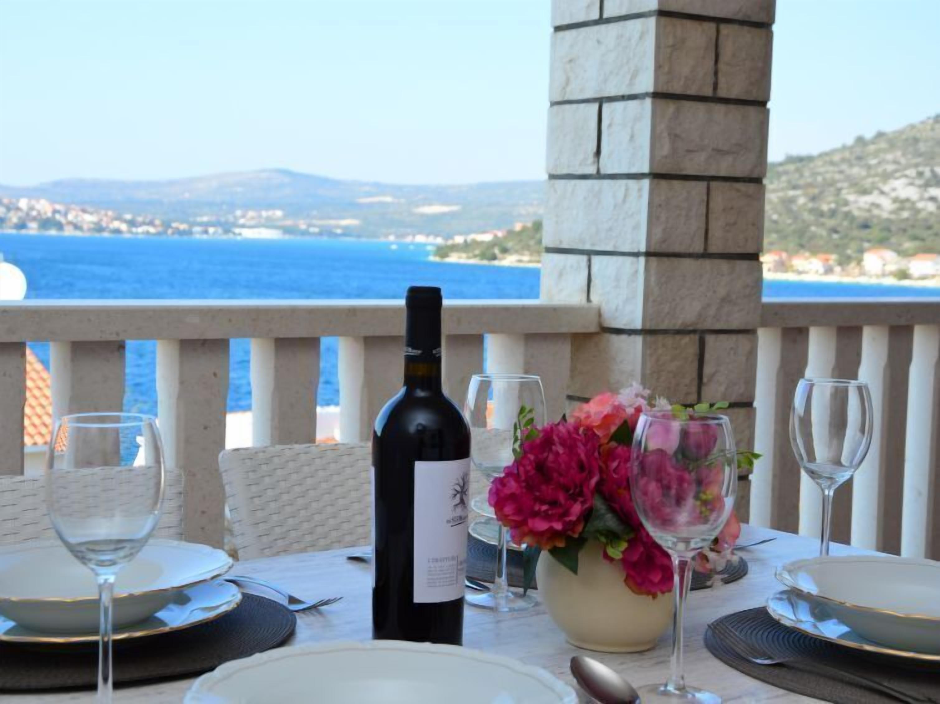 A table set for dining with a bottle of wine, glasses, and a vase of flowers, overlooking a coastal view.