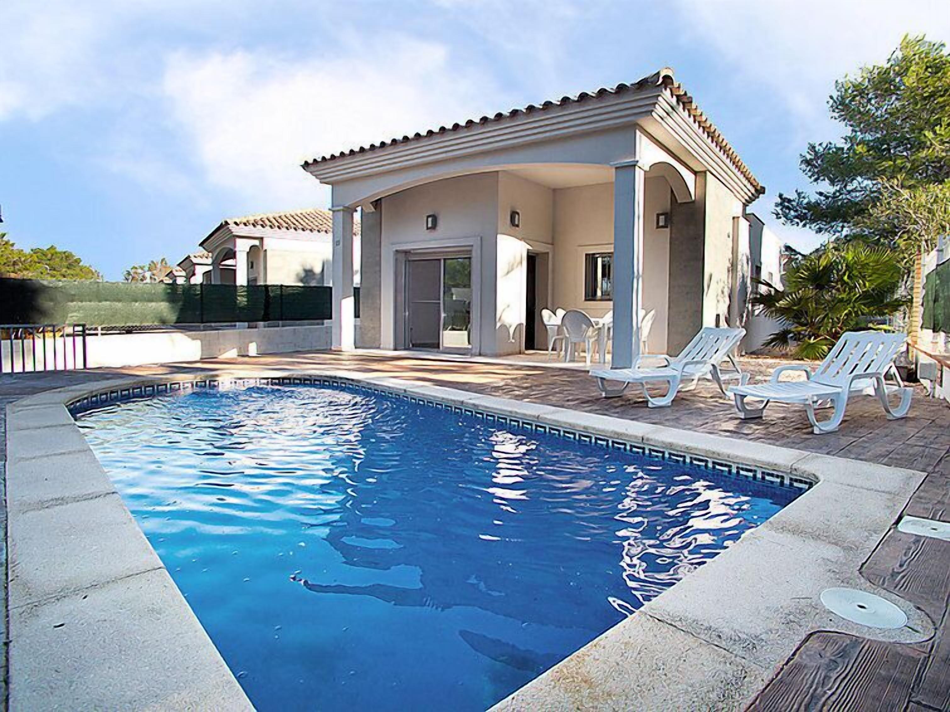 A swimming pool with a tiled deck, white lounge chairs, and a covered patio area with tables and chairs.