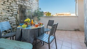 Un pequeño comedor al aire libre con muros de piedra, una mesa dispuesta con diversas frutas y una jarra de jugo de naranja, y vistas de edificios a lo lejos.