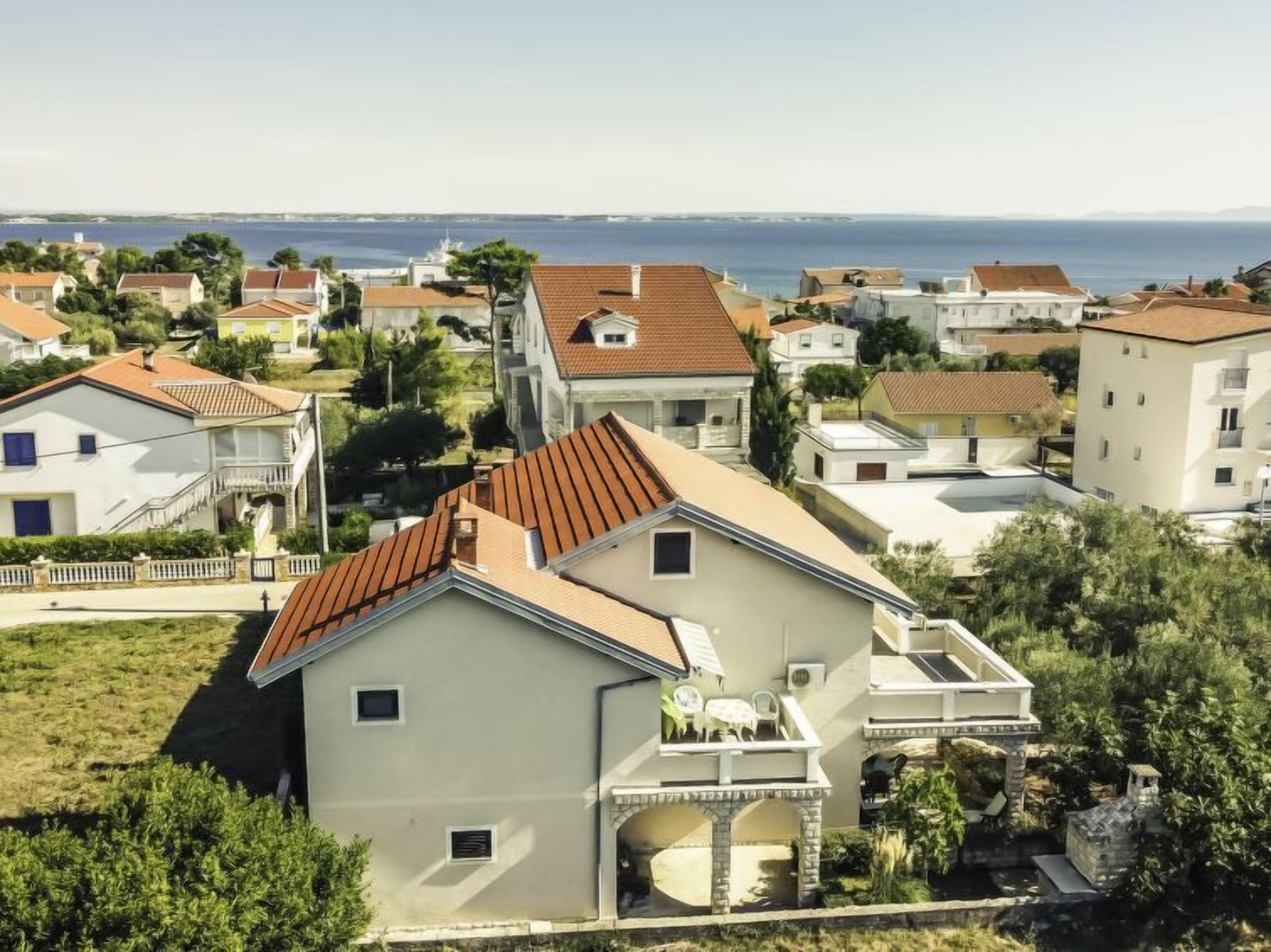 A residential area with houses featuring red-tiled roofs and light-colored walls, surrounded by greenery and a view of the sea.