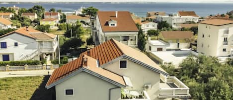 A residential area with houses featuring red-tiled roofs and light-colored walls, surrounded by greenery and a view of the sea.