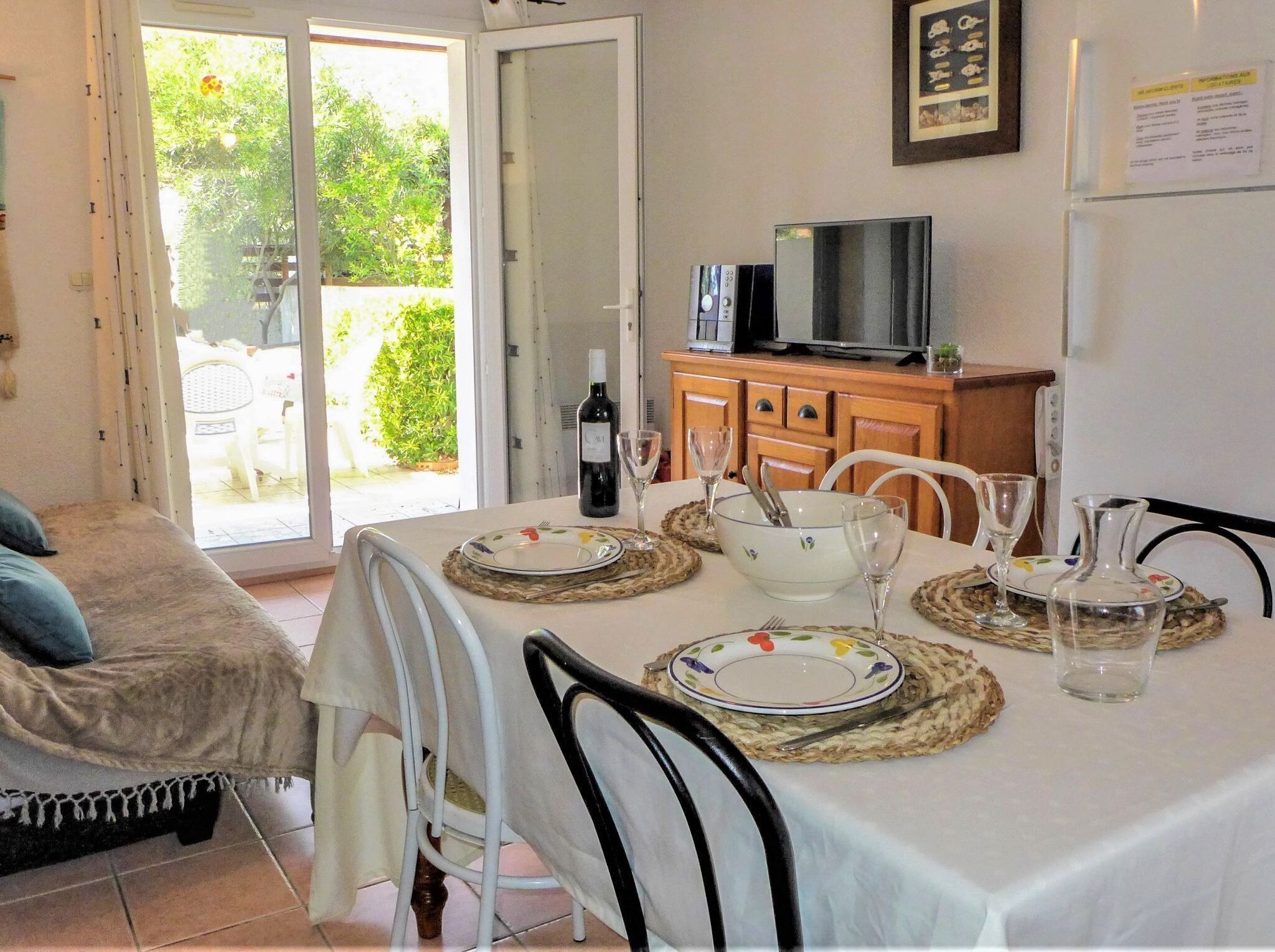 Un coin repas avec une table dressée pour deux personnes, un téléviseur posé sur un meuble en bois et une porte donnant sur un balcon.
