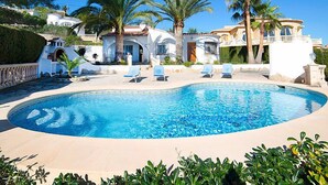 Une piscine entourée de chaises longues, de palmiers et, au loin, de villas de style méditerranéen.