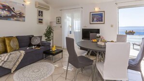 A modern living room with a sofa, TV, dining table, and chairs. There are framed pictures on the walls and a large window with a view of the sea.