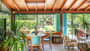 Une pièce spacieuse avec un plafond haut, des poutres en bois et de grandes fenêtres donnant sur un jardin luxuriant.