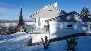 Una casa moderna con balcón de vidrio, rodeada por un paisaje nevado y árboles.