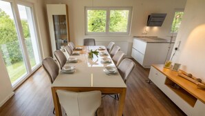 Un comedor moderno con una mesa larga dispuesta para seis personas, rodeada de sillas. Hay una zona de cocina con una encimera y un estante con adornos.