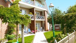 Un edificio residenziale a due piani con facciata in mattoni, un balcone con ringhiera in vetro e un piccolo giardino con prato e altalena.