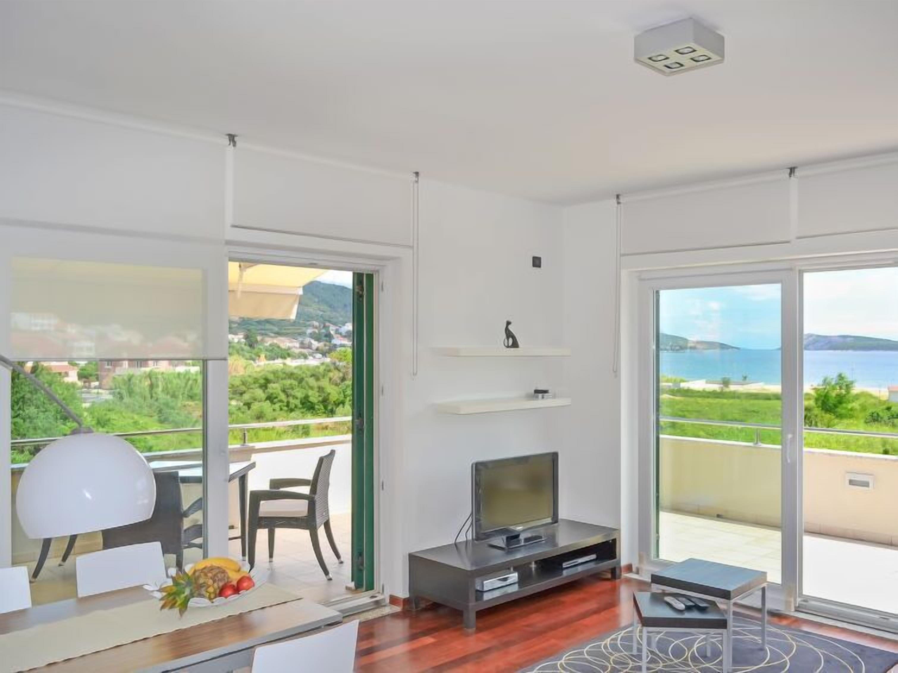 Una sala de estar moderna con televisor, un comedor con mesa y sillas, y ventanas amplias con vistas al mar y al verdor.