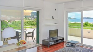 A modern living room with a TV, a dining area with a table and chairs, and large windows offering a view of the sea and greenery. - Jadranka by Interhome (Rab/Rab)