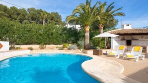 Una piscina amb voreja corba, envoltada per un solàrium de rajoles blanques. Hi ha gandules amb coixins grocs i un parasol blanc. Palmeres i vegetació abundant es veuen al fons.