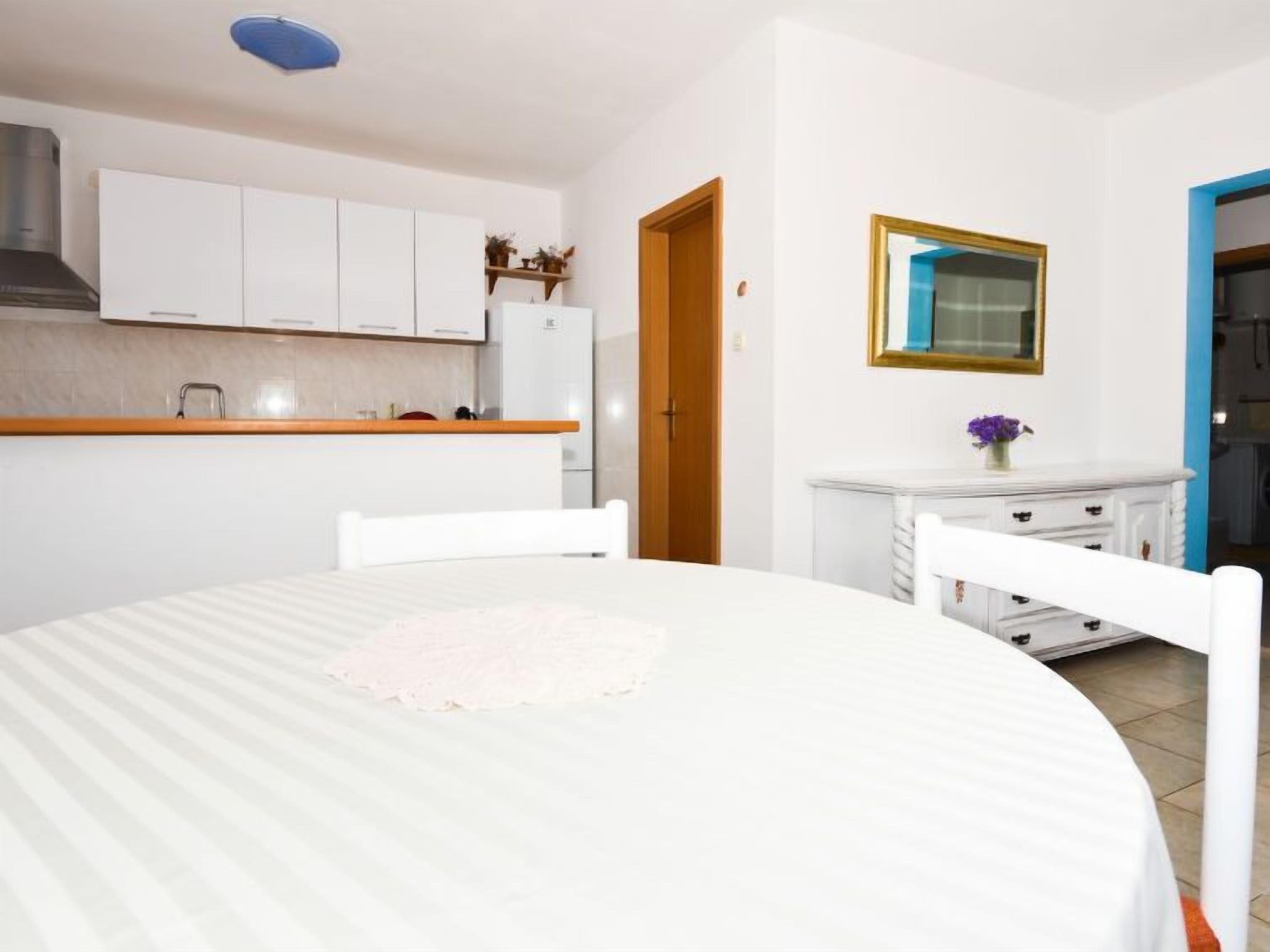 A small, clean room with a round dining table, white chairs, a small kitchen area with cabinets, and a door leading to another room.