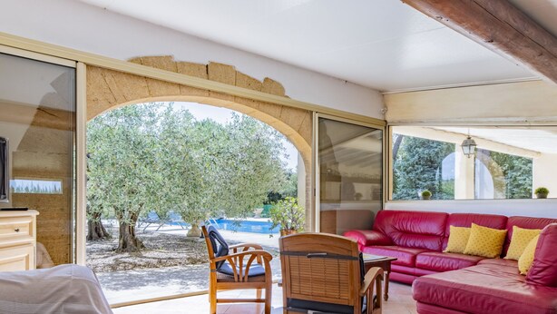 Una habitación con una cama, una silla de madera, un sofá de cuero rojo y vistas a una piscina exterior y árboles.