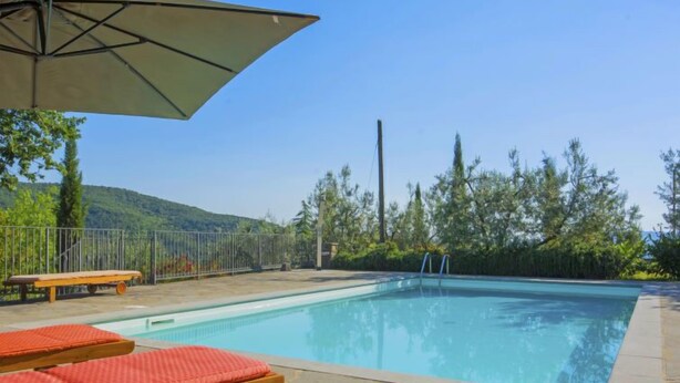 A swimming pool with lounge chairs and an umbrella, surrounded by a fence and trees.