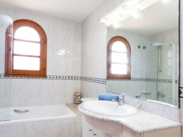 Un baño con bañera, lavabo con encimera de mármol y un espejo grande sobre el lavabo. Se ven dos ventanas con marcos de madera y un detalle decorativo en la pared.