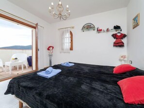 A bedroom with a black bedspread, a red pillow, a white chair, and a small table with a vase.