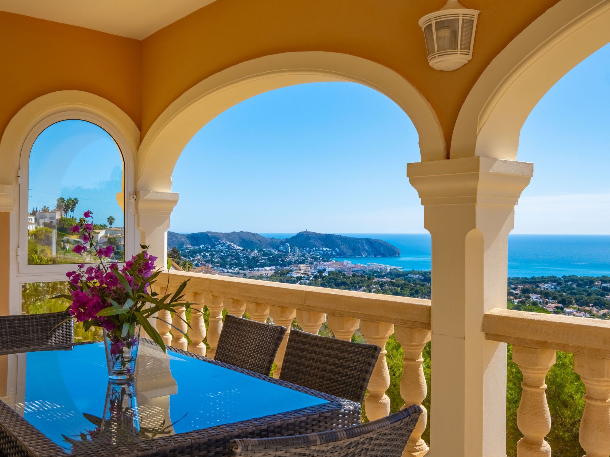 Un balcón con una mesa y sillas, un jarrón con flores y vistas al mar y al pueblo.