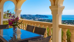 A balcony with a table and chairs, a vase with flowers, and a view of the sea and town. - La Perla by Interhome (Moraira)