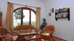 A cozy room with wicker furniture, a large window with curtains, a bookshelf, and a potted plant. - Balcon al Mar by Interhome (Jávea)