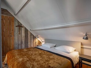 Ein Schlafzimmer mit schrägen Wänden, Holzwandpaneelen an einer Wand, einem Bett mit weißer Bettwäsche und einer Nachttischlampe.