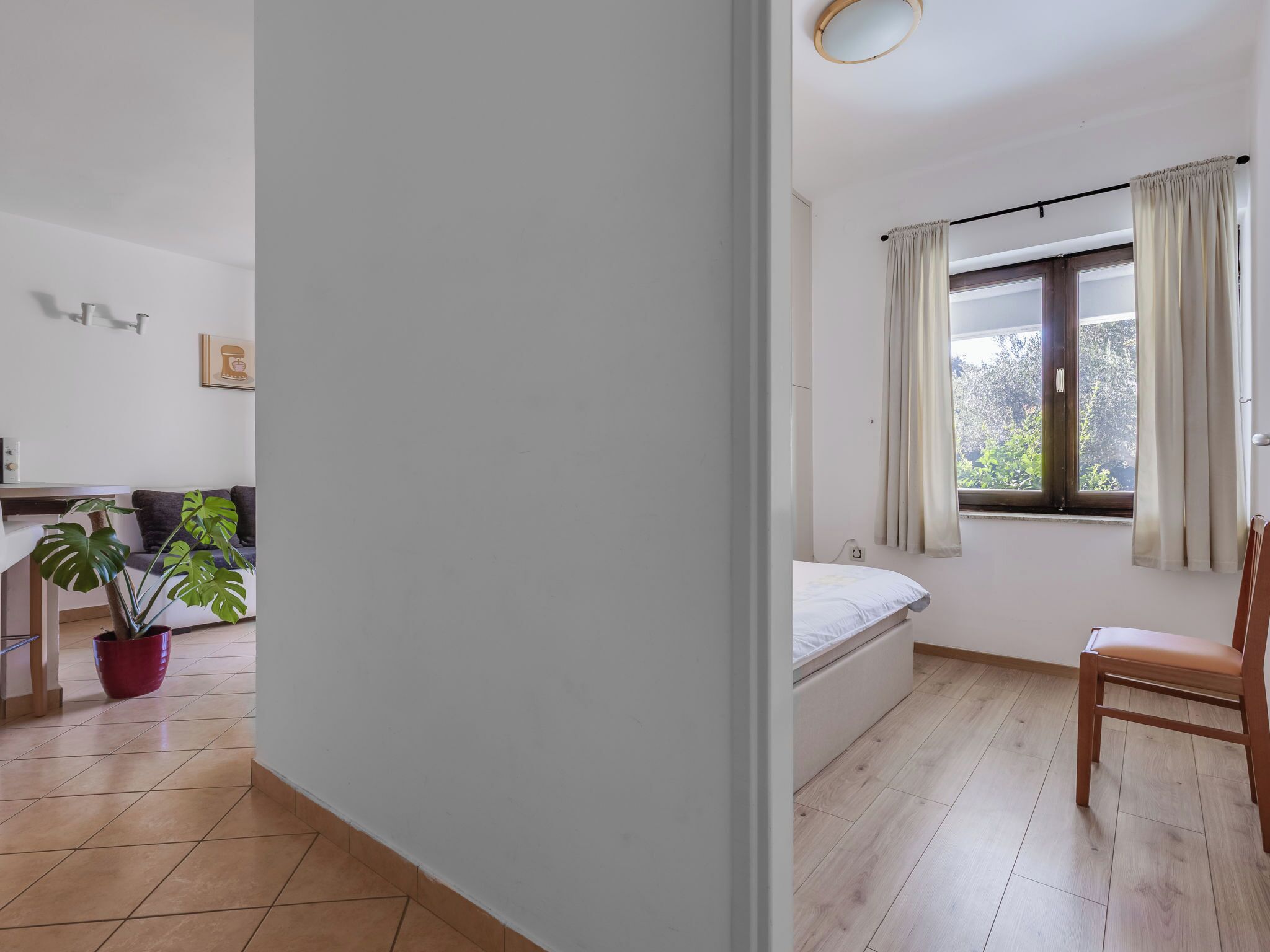 Een slaapkamer met een enkel bed, een stoel, een raam met gordijnen, een kamerplant en uitzicht op groen buiten.
