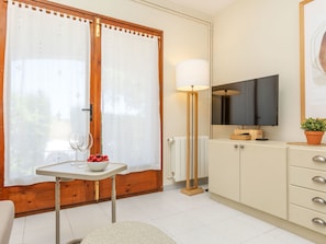Una habitación con puerta de madera, una mesa blanca con un frutero y copas de vino, una lámpara de pie, un televisor sobre un mueble y una planta en maceta.