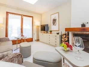 Una sala moderna con un sofá, otomanas, una mesa de comedor con un frutero y copas de vino, una chimenea y un televisor sobre un mueble.