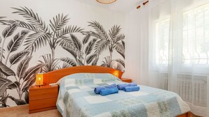 A bedroom with a bed, wooden nightstands, a wall with a leaf pattern, a hanging light fixture, and a window with sheer curtains. - Vacation home Antem by Interhome (Lloret de Mar)