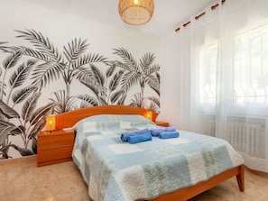 A bedroom with a bed, wooden nightstands, a wall with a leaf pattern, a hanging light fixture, and a window with sheer curtains. - Vacation home Antem by Interhome (Lloret de Mar)