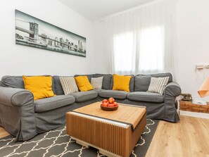 Moderni olohuone, jossa on harmaa kulmasohva, keltaiset koristetyynyt, puinen sohvapöytä, jossa on omenoita, ja seinällä kaupunkimaisemaa esittävä taulu.