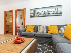 Ett vardagsrum med en grå soffa, gula kuddar, ett träbord med äpplen och en tavla som föreställer en stadsbild på väggen.