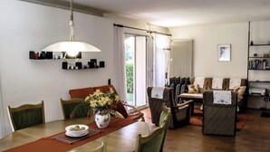 Un coin repas avec une table en bois, des chaises et un vase de fleurs. On aperçoit un canapé et une bibliothèque à l’arrière-plan.