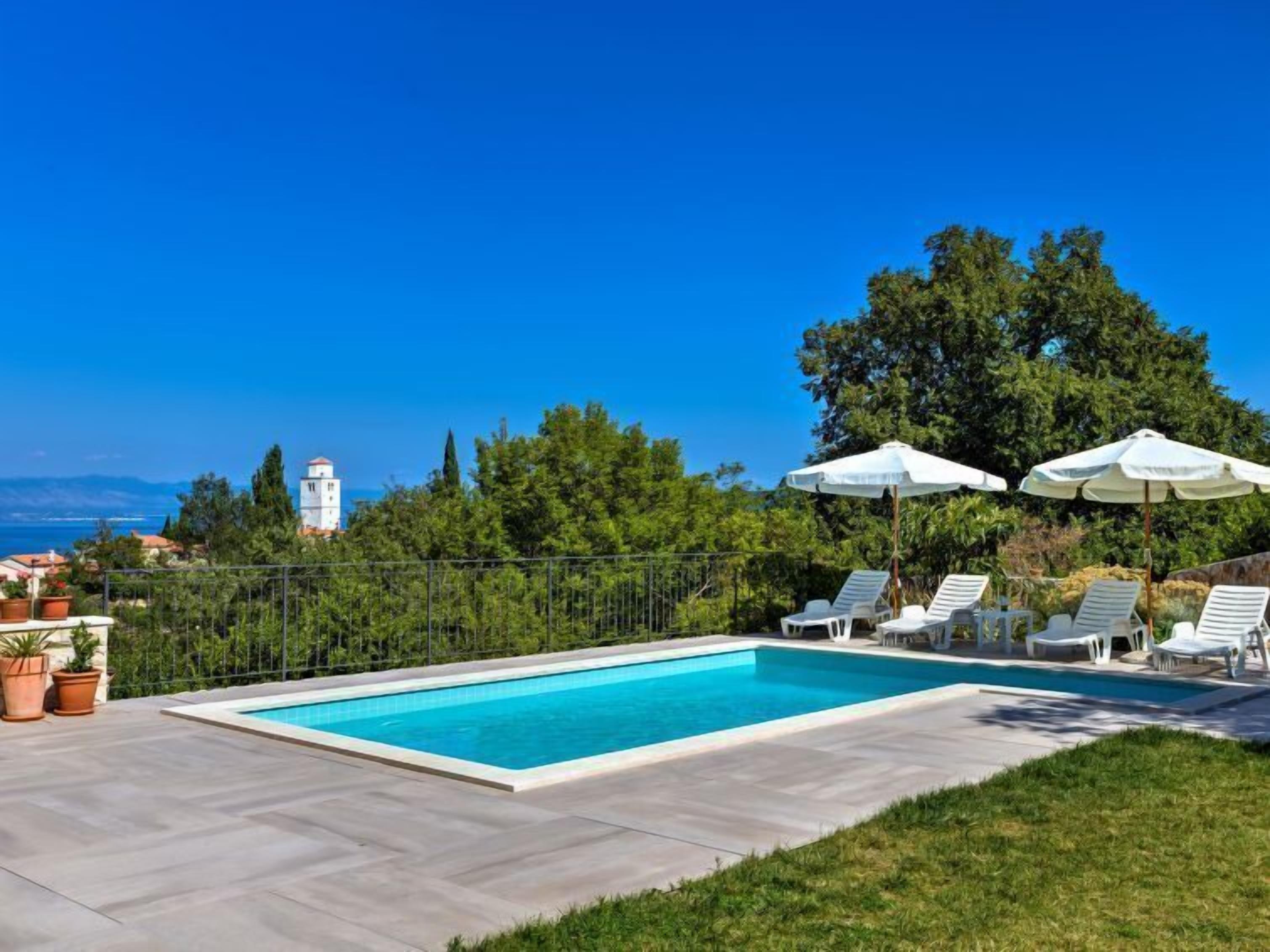 A rectangular swimming pool with white lounge chairs and umbrellas, surrounded by a paved patio and a metal fence.
